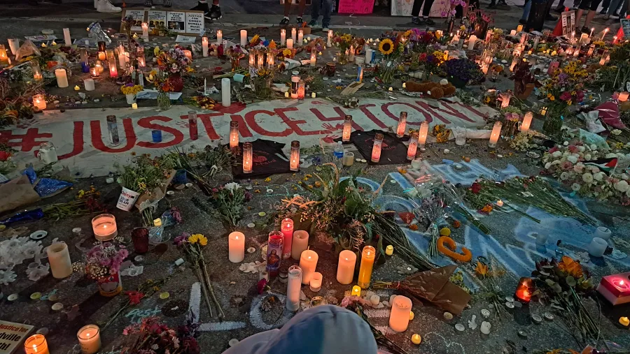 A photo of a vigil in the aftermath of the shooting of Corey Ruiz shows a number of lit candles arrancged on the ground, alongside flowers, chalk drawings, and protest signs.